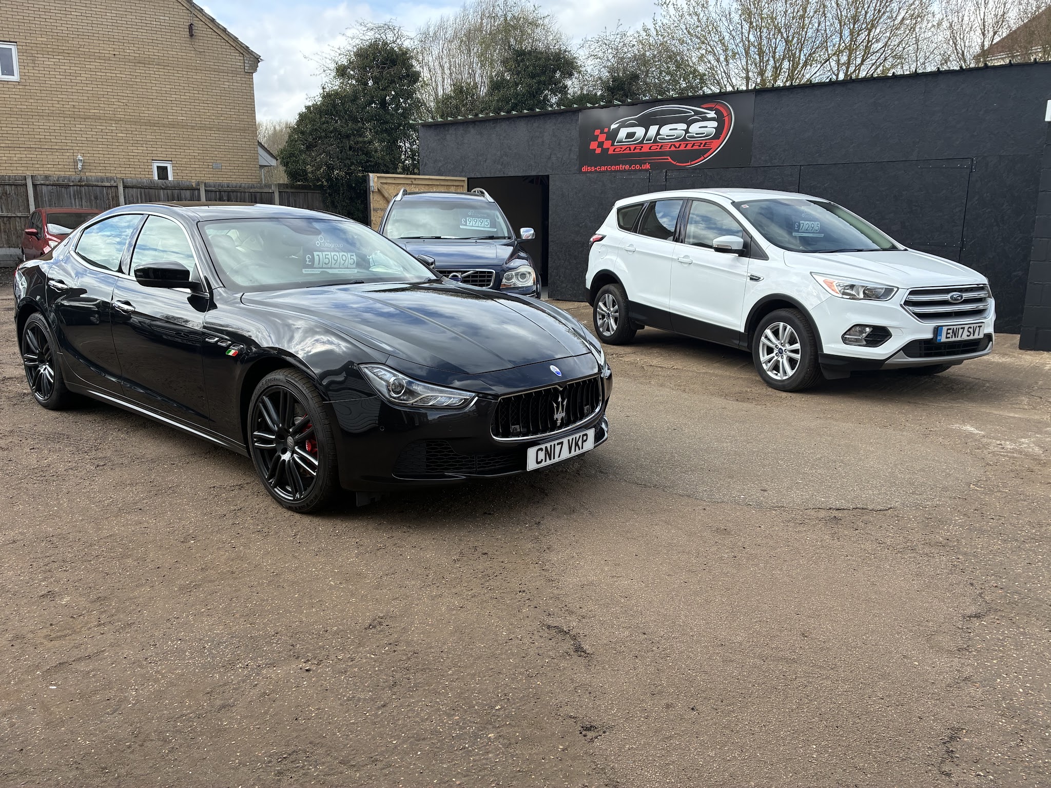 A black Maserati Ghibli and a white Ford Kuga on the Diss Car Centre forecourt, in front of the red and black sign