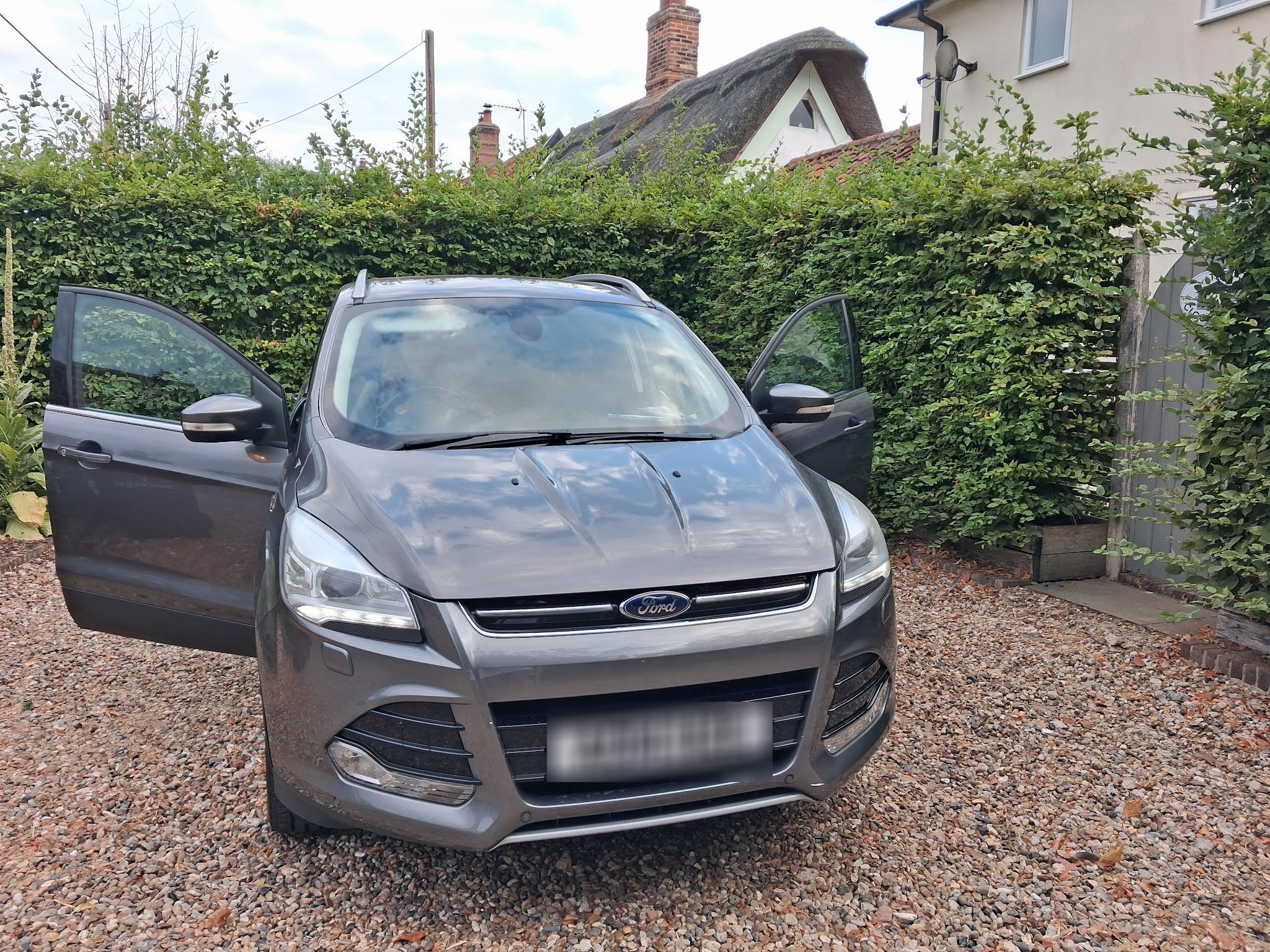 The grey Ford Kuga delivered to Keith and Julia at their holiday cottage, doors open, in front of a hedge